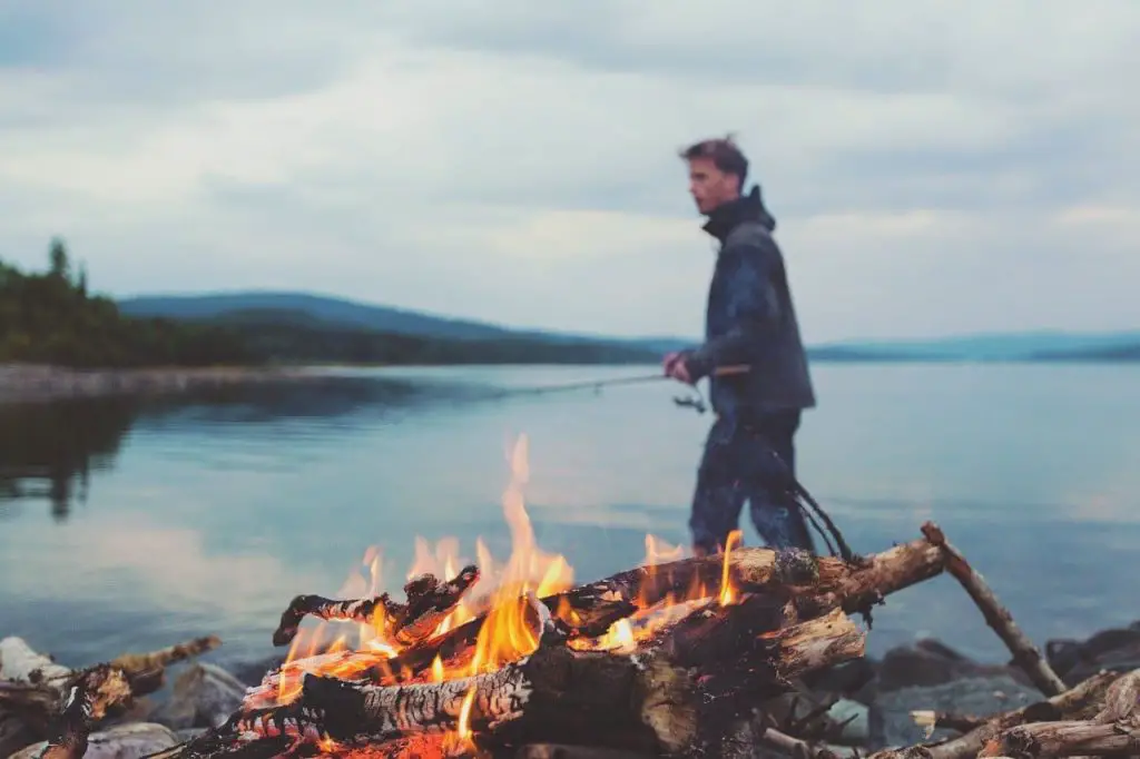 a guy camping besides a lake and a bonfire